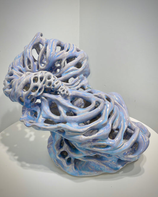An abstract sculptural object with a glossy, organic, coral-like or bone-like structure in pale blue and white, displayed on a white surface against a neutral light gray background.